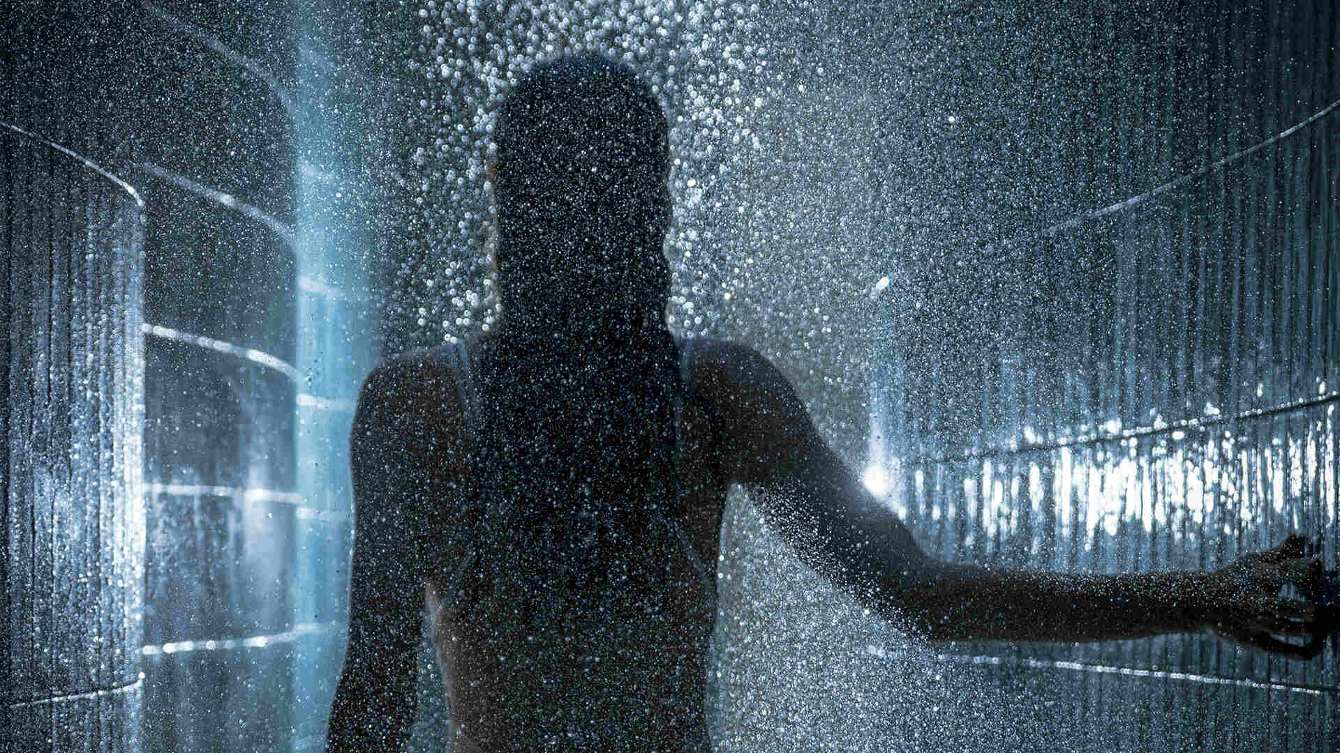 Silhouette of a woman standing under cascading water in a luxurious rainforest-themed spa shower. Surrounded by dark tiled walls and soft ambient lighting, the scene evokes a sense of deep relaxation and immersive wellness.